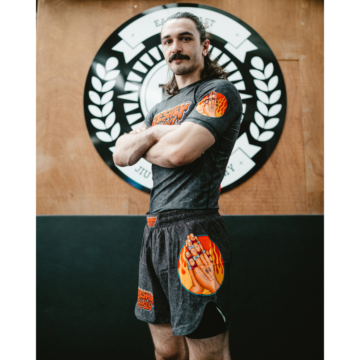 A man with a mustache and long hair stands with arms crossed, wearing a dark gray athletic t-shirt and matching shorts with orange and yellow graphic designs. He is standing in front of a wooden wall with a circular black and white logo.