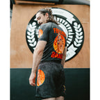 A man wearing a black graphic t-shirt and matching shorts with orange and yellow flame designs, standing in front of a wooden wall with a circular logo.