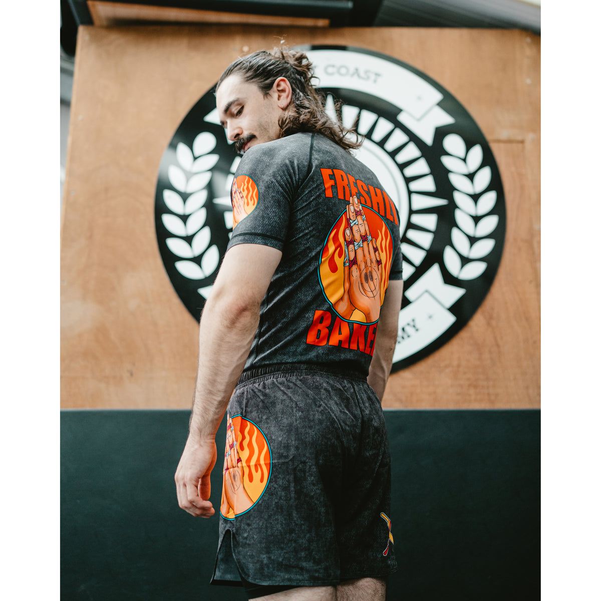 A man wearing a black graphic t-shirt and matching shorts with orange and yellow flame designs, standing in front of a wooden wall with a circular logo.
