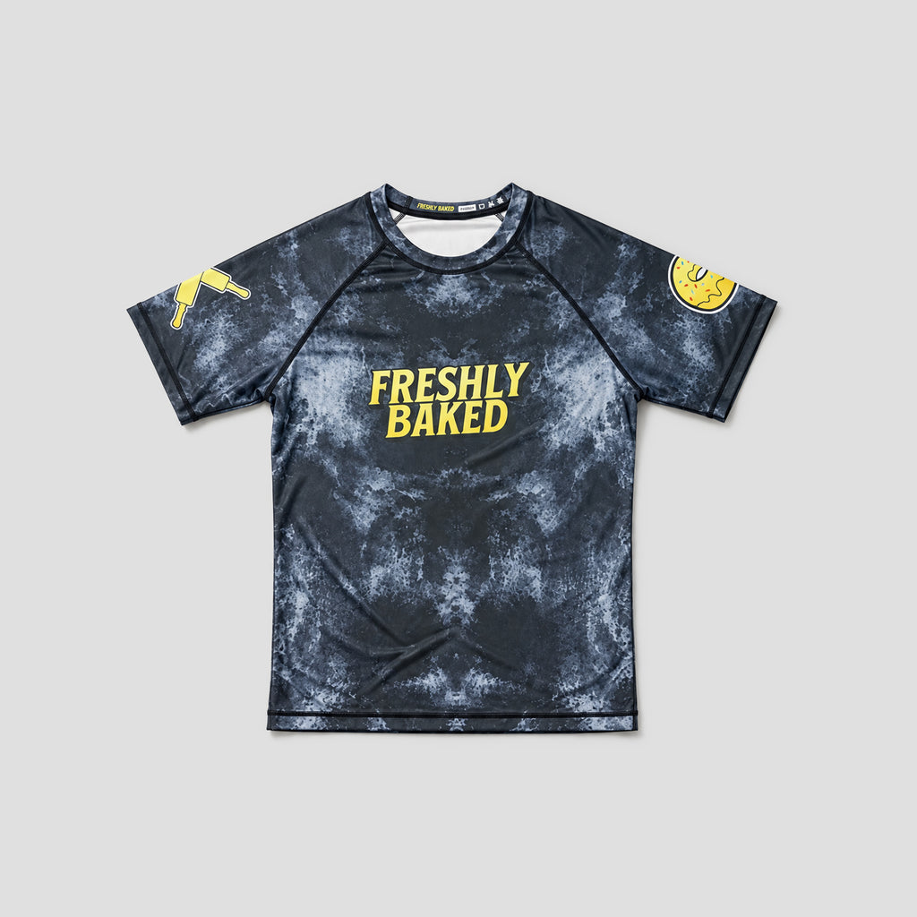 A short-sleeve athletic t-shirt with a dark gray and black mottled pattern, featuring the words 'FRESHLY BAKED' in yellow on the chest, a gold donut face on the right sleeve, and a gold graphic on the left sleeve, displayed against a light gray background.