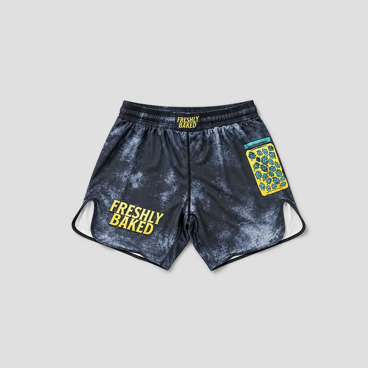 A pair of athletic shorts with a dark gray and white marble pattern, featuring 'FRESHLY BAKED' printed in yellow on the left leg and a yellow and blue patterned patch on the right leg.