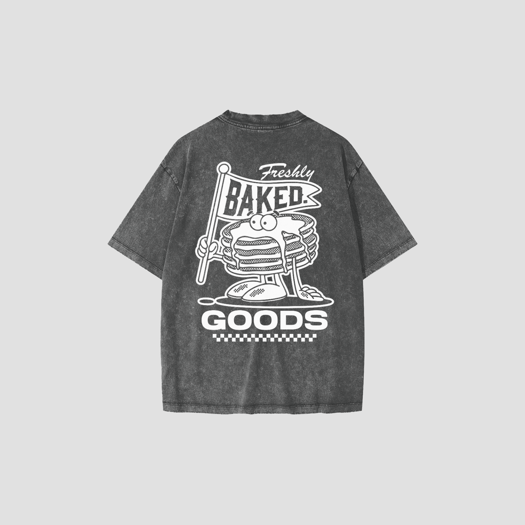 A dark gray acid-wash t-shirt shown from the back, featuring a white graphic print of a stack of pancakes with a flag that reads 'Freshly Baked' and the words 'GOODS' below.