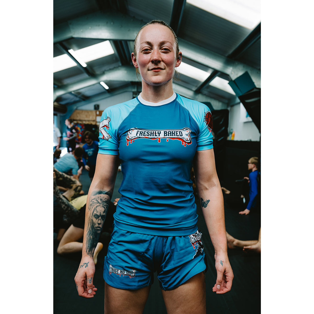 A woman standing in a gym wearing a blue and light blue short-sleeve athletic jersey with 'Freshly Baked' printed across the chest, paired with matching blue shorts.