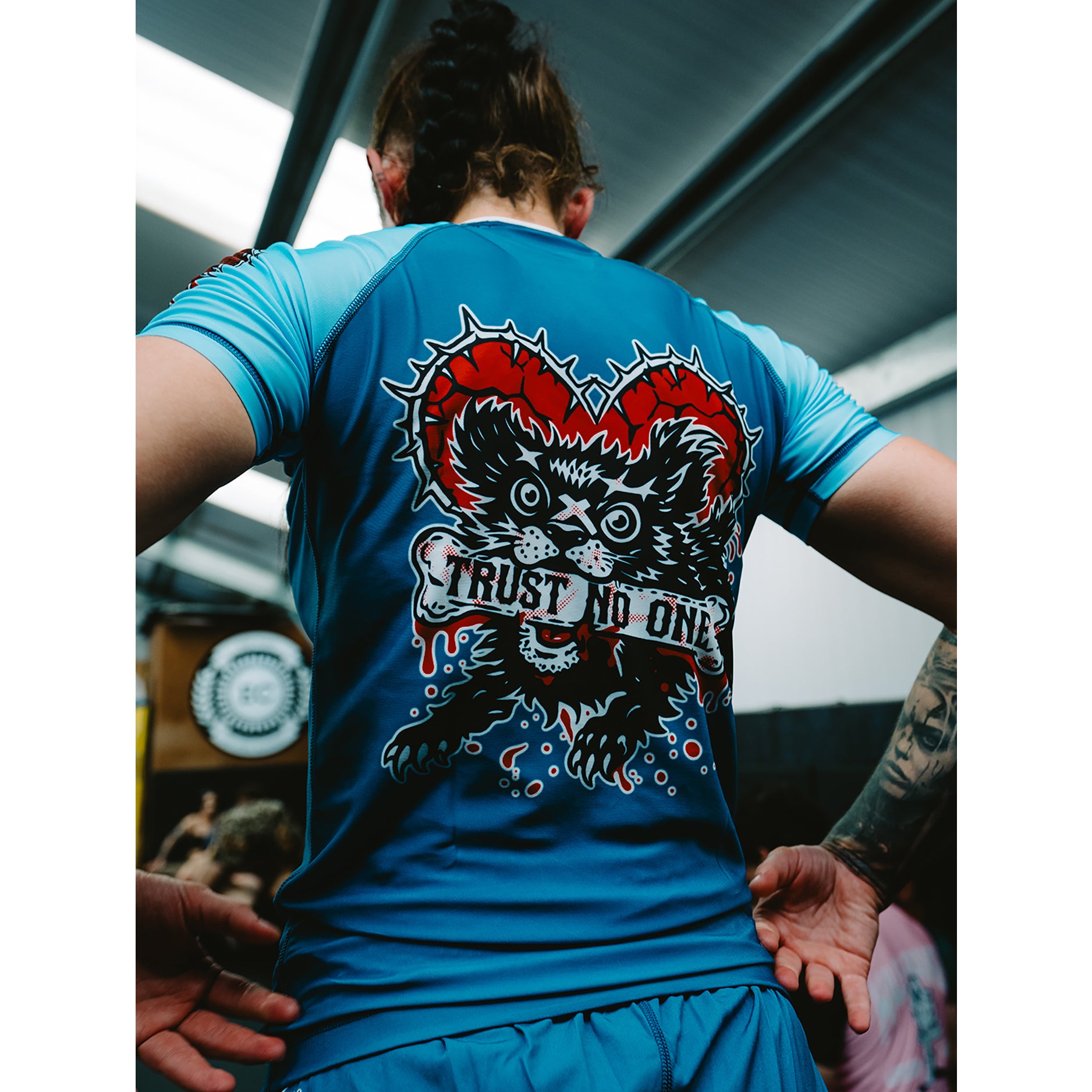 A person viewed from behind wearing a bright blue athletic T-shirt with a large back graphic showing a black cat inside a red heart and the words “TRUST NO ONE.”.