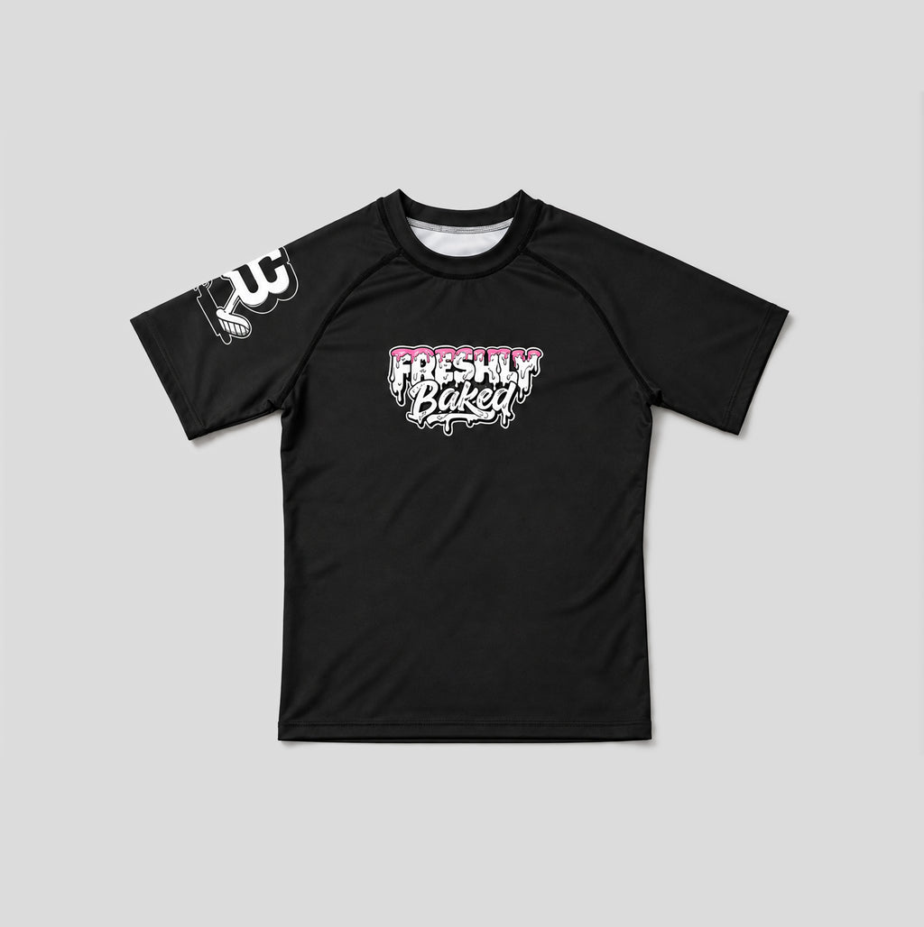 A black short-sleeve rash guard with a 'Freshly Baked' graphic in pink and white on the chest, and a white graphic on the right sleeve, displayed flat against a light gray background.