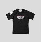 A black short-sleeve rash guard with a 'Freshly Baked' graphic in pink and white on the chest, and a white graphic on the right sleeve, displayed flat against a light gray background.