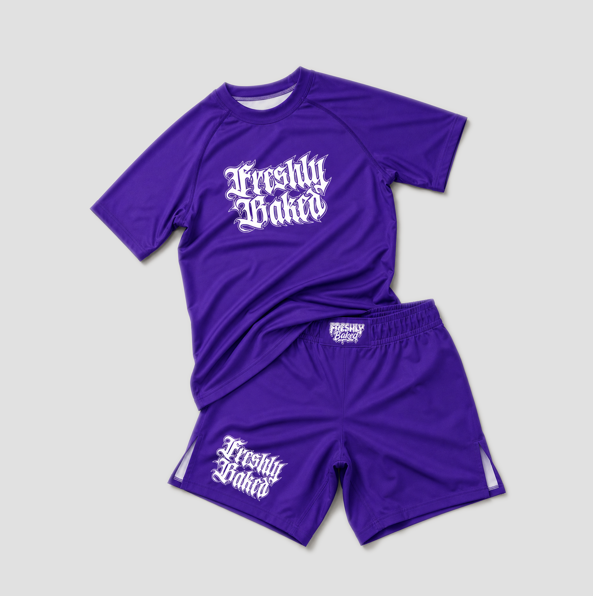 A two-piece athletic set consisting of a purple short-sleeve t-shirt and matching shorts, both featuring the text 'Freshly Baked' in a white gothic font. The t-shirt has a larger chest graphic, while the shorts have a smaller one on the left leg.