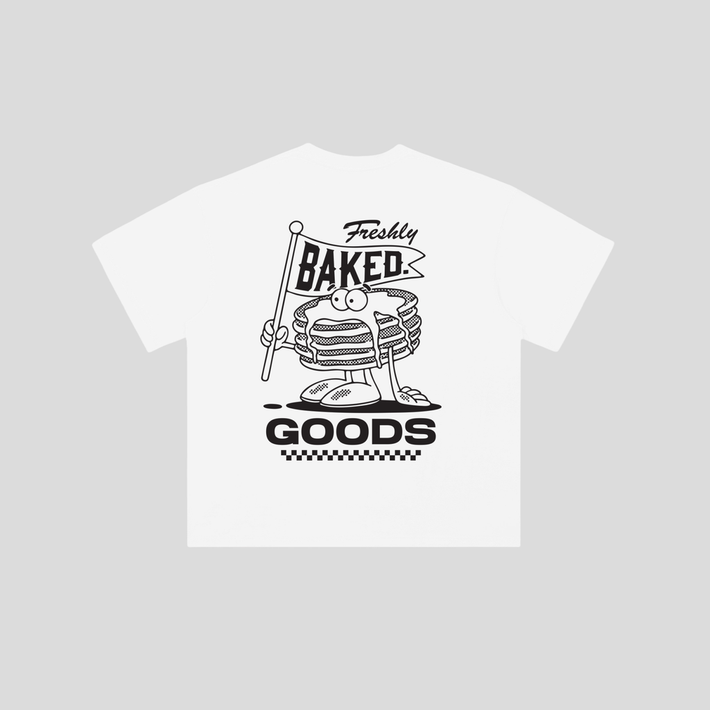 A white short-sleeve t-shirt with a black graphic print of a cartoon stack of pancakes holding a 'Freshly Baked Goods' sign, displayed on a solid light gray background.