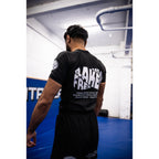 A man viewed from behind wearing a black athletic t-shirt with 'FRESHLY BAKED' printed in bold white letters on the back, inside a gym with blue mats on the floor.