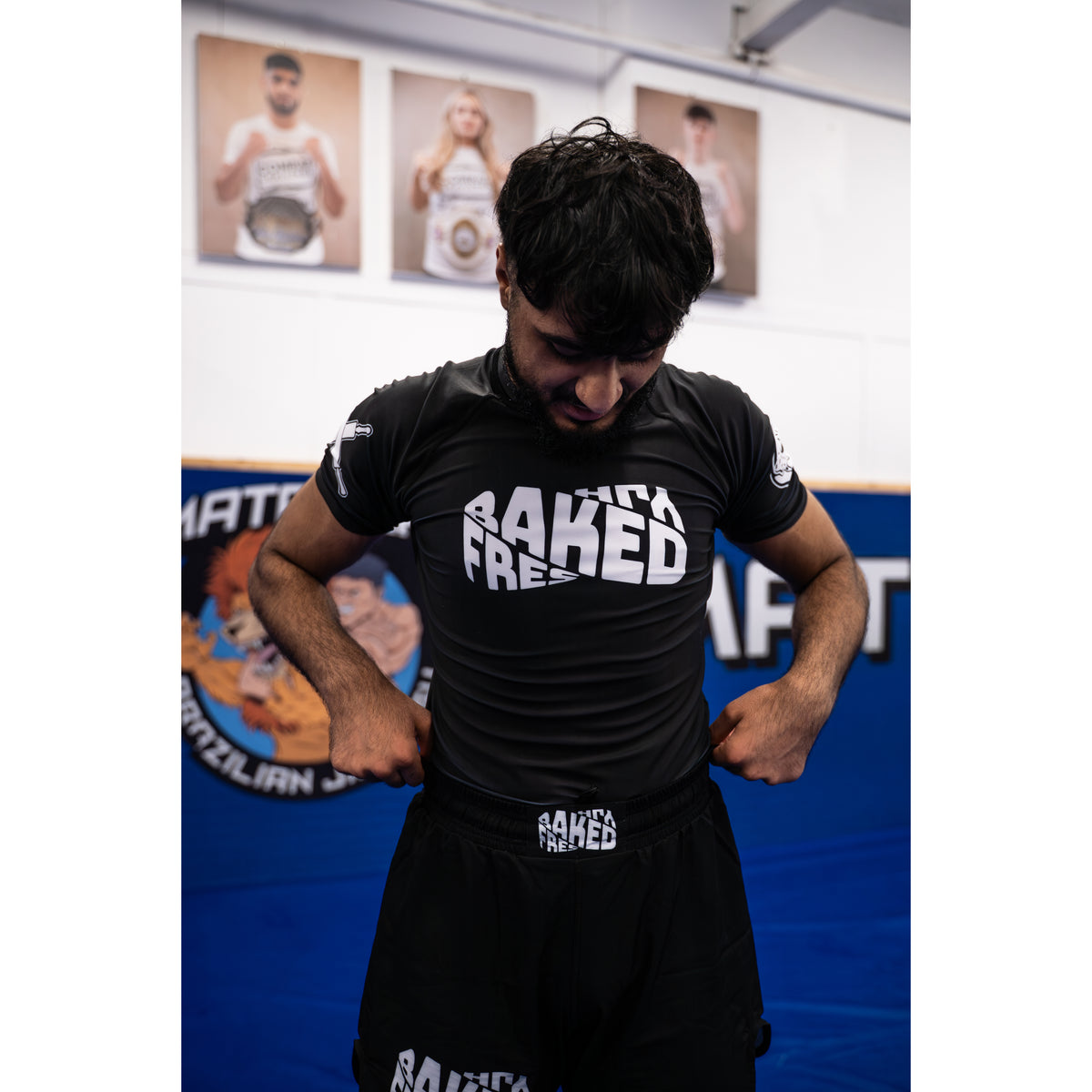 A man wearing a black athletic rash guard and matching black shorts with white 'FRESHLY BAKED' branding, standing in a gym with a blue mat background.