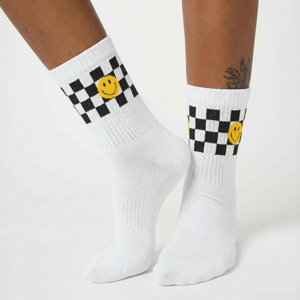 White crew socks with a black and white checkered band, a yellow smiley face, and ribbed knit detailing, shown on a model's feet against a light gray background.