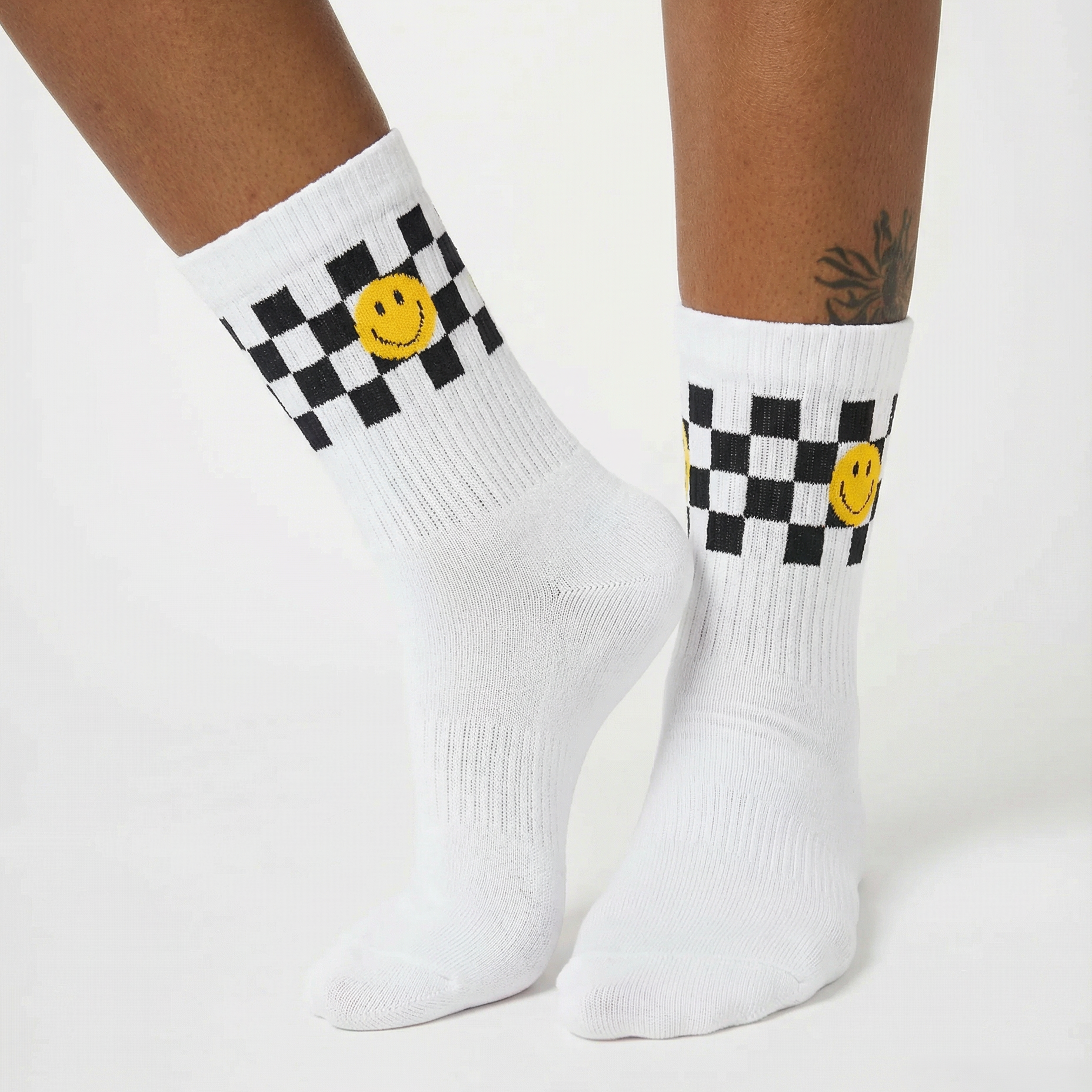 White crew socks with a black and white checkered band, a yellow smiley face, and ribbed knit detailing, shown on a model's feet against a light gray background.