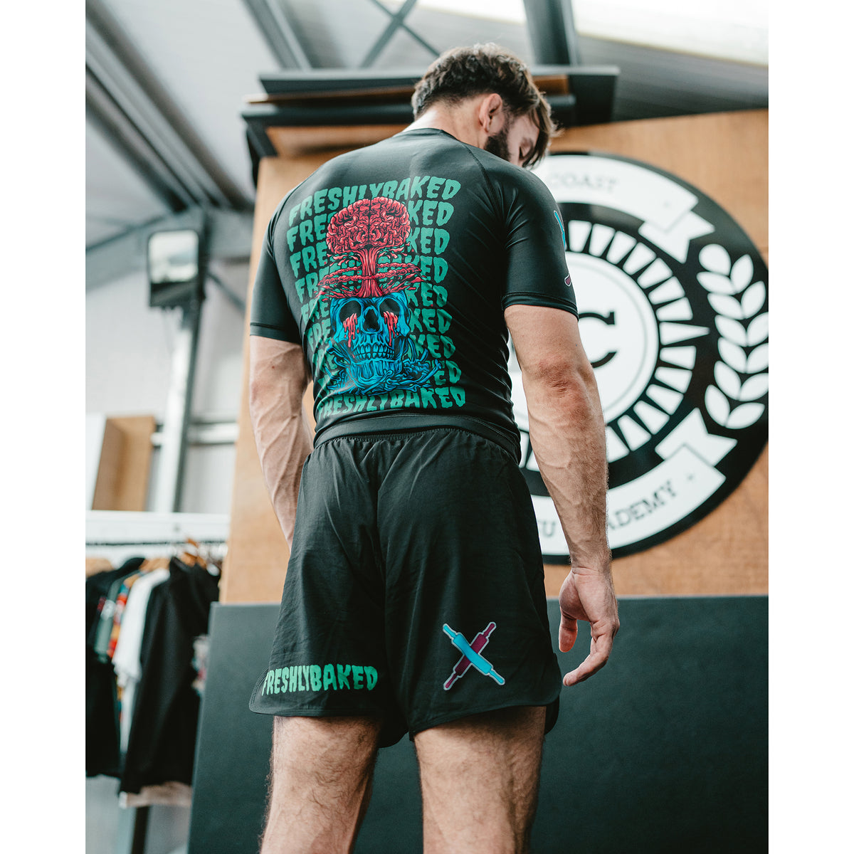A man wearing a black Freshly Baked rash guard and matching black swim shorts, viewed from behind in an indoor gym or studio setting. The rash guard features a large back graphic with a red tree, blue skull, and teal text, while the shorts have a small teal graphic near the hem.