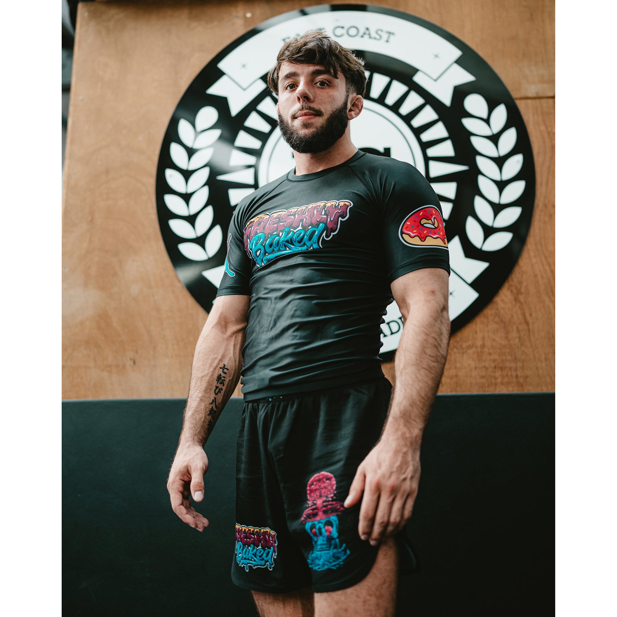 A man wearing a black short-sleeve athletic t-shirt and matching black shorts with colorful graphic prints, standing in front of a wooden wall with a large circular black-and-white logo.