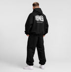 A man wearing a black hoodie and sweatpants, shown from the back against a light gray studio background. The hoodie features a large white graphic text print on the back reading 'Freshly Baked'.