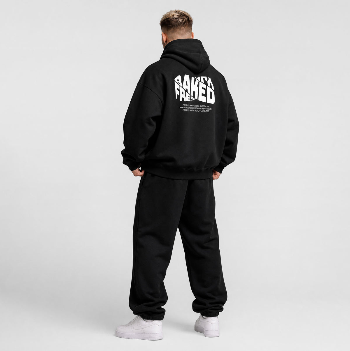 A man wearing a black hoodie and sweatpants, shown from the back against a light gray studio background. The hoodie features a large white graphic text print on the back reading 'Freshly Baked'.