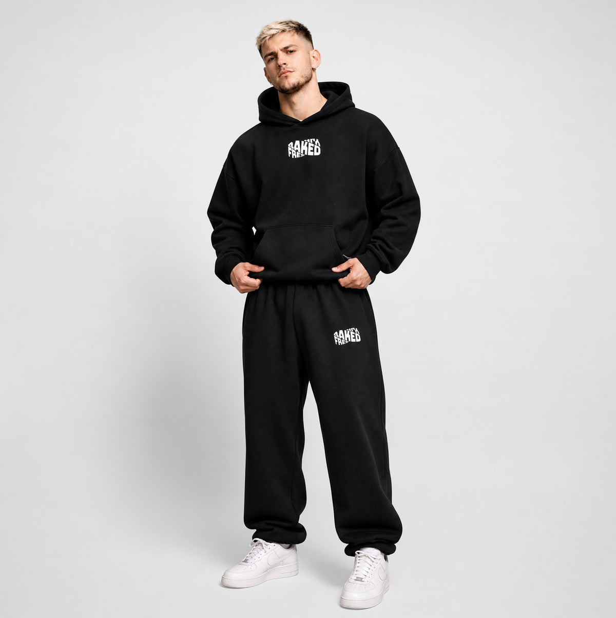 A man modeling a black two-piece hoodie and sweatpants set with white 'Freshly Baked' text graphics on the chest and thigh, paired with white sneakers, against a light gray studio background.