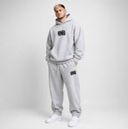 A man modeling a light gray heathered hoodie and matching sweatpants set with black 'FRESHLY BAKED' graphics, standing against a light gray studio background.