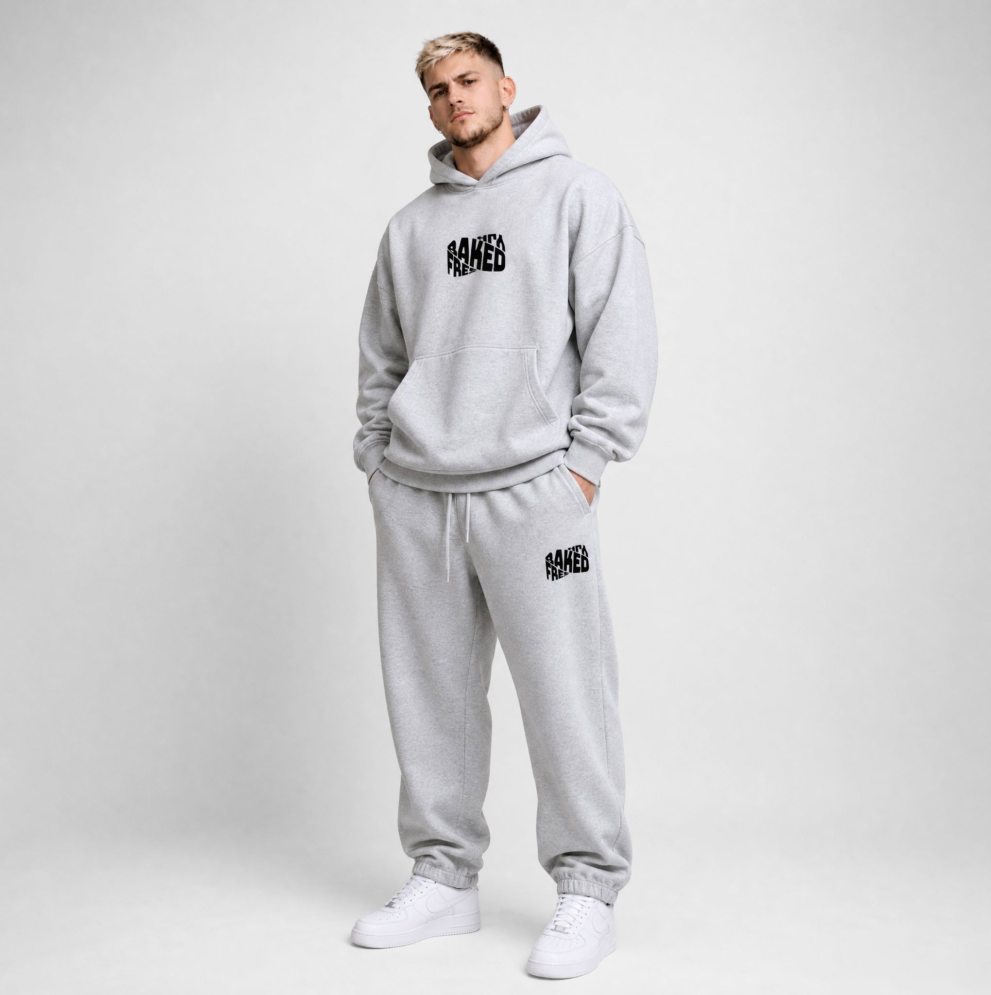 A man modeling a light gray heathered hoodie and matching sweatpants set with black 'FRESHLY BAKED' graphics, standing against a light gray studio background.