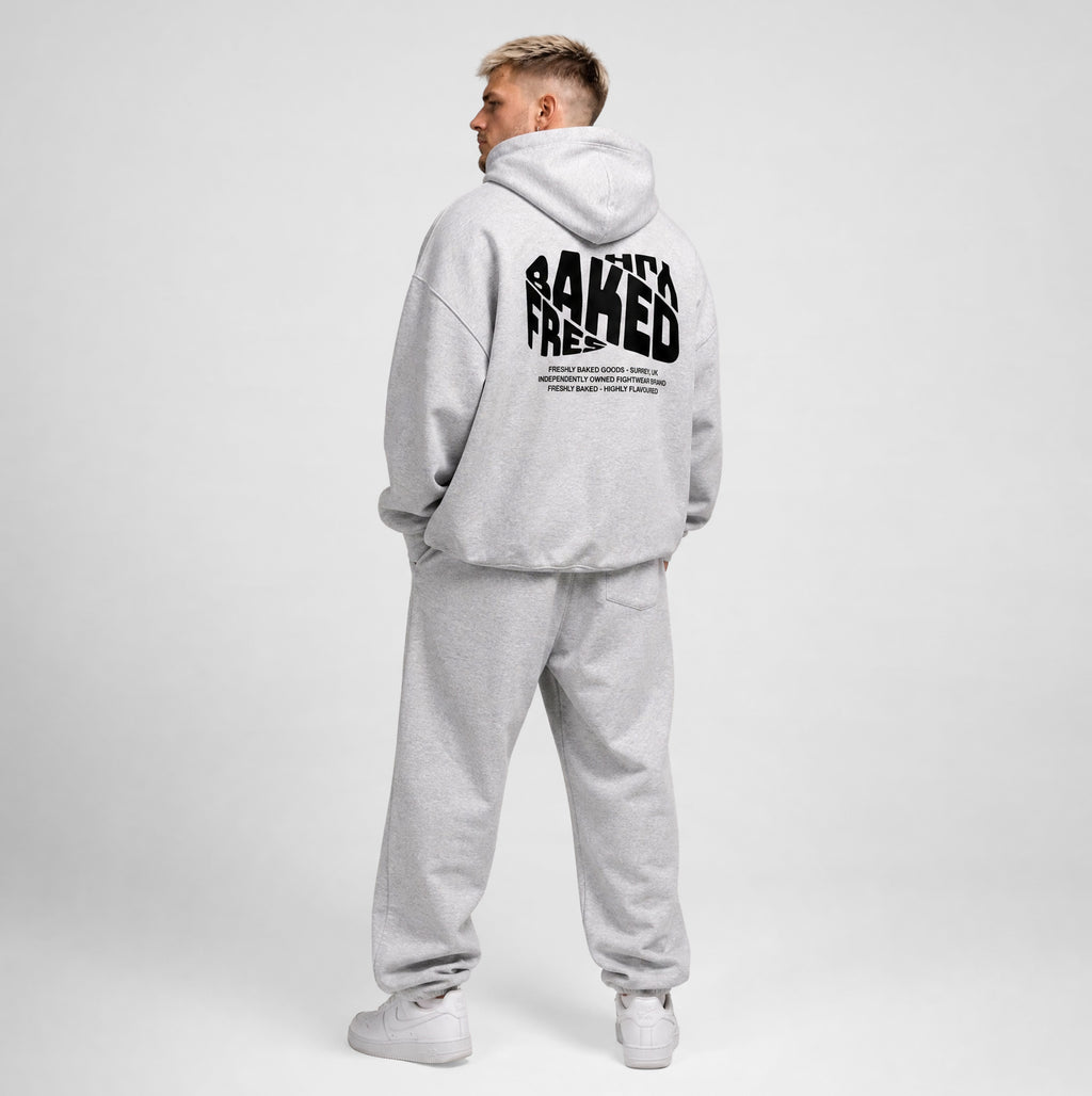 A man wearing a light gray heathered hoodie and matching sweatpants, shown from the back. The hoodie features a large black graphic text print on the back reading 'BAKED'.