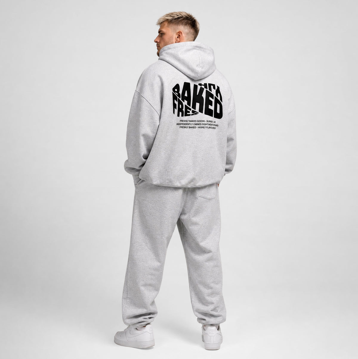 A man wearing a light gray heathered hoodie and matching sweatpants, shown from the back. The hoodie features a large black graphic text print on the back reading 'BAKED'.