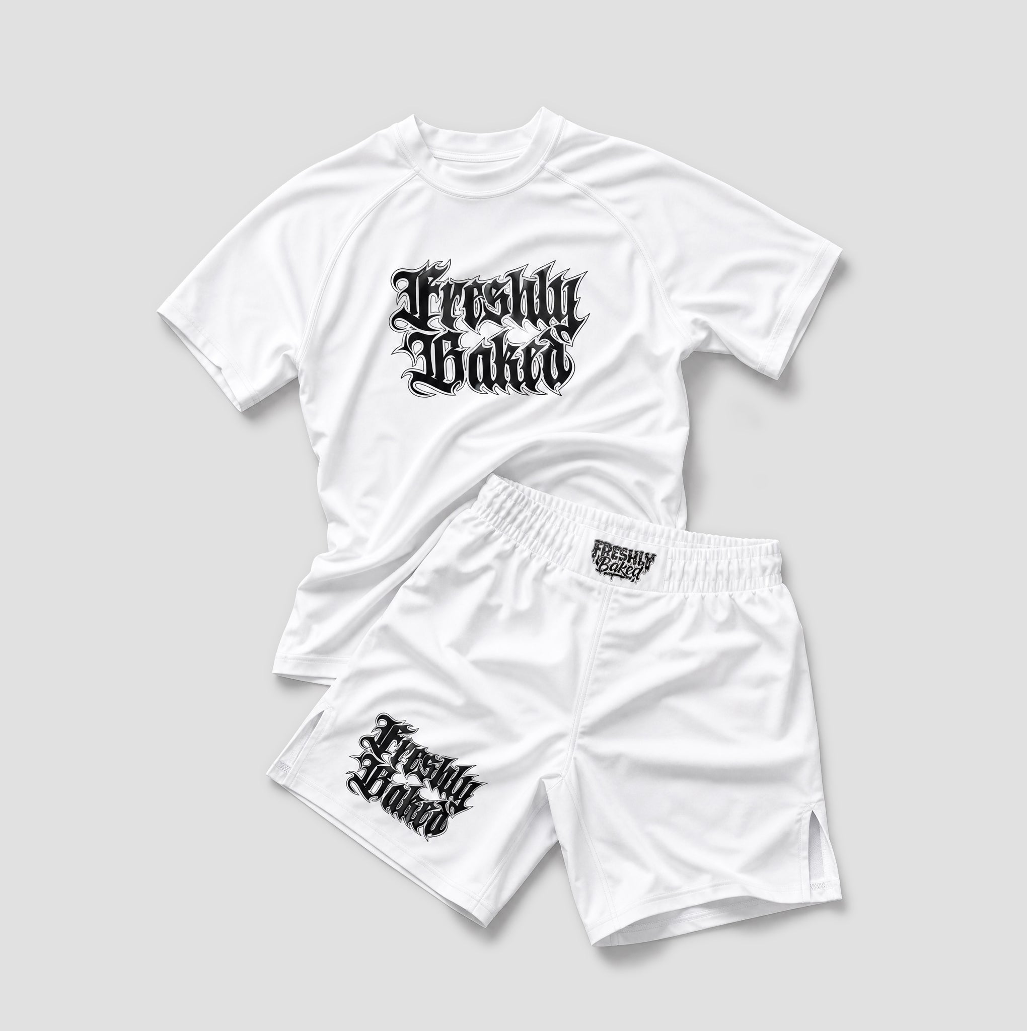 A white athletic two-piece set consisting of a short-sleeve raglan t-shirt and matching shorts, both featuring a black gothic-style 'Freshly Baked' graphic. The t-shirt is laid flat on top of the shorts against a neutral light gray background.