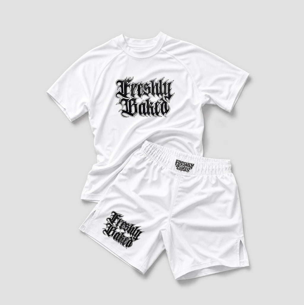 A white athletic two-piece set consisting of a short-sleeve raglan t-shirt and matching shorts, both featuring a black gothic-style 'Freshly Baked' graphic. The t-shirt is laid flat on top of the shorts against a neutral light gray background.