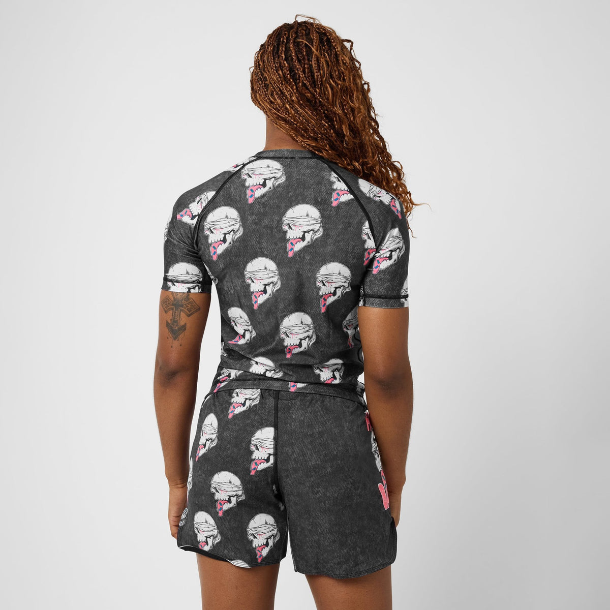 A woman shown from the back wearing a dark gray short-sleeve rash guard and matching shorts with an all-over football helmet print in white, pink, and black, against a light gray studio background.