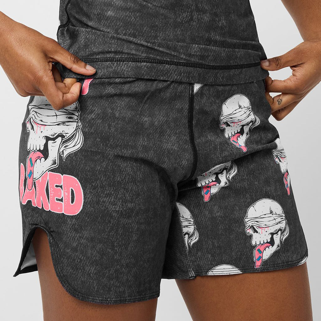 Close-up of a person wearing dark gray heathered shorts with a repeating skull-and-mouth graphic and large pink 'BAKED' text on the left leg, shown against a light gray studio background.