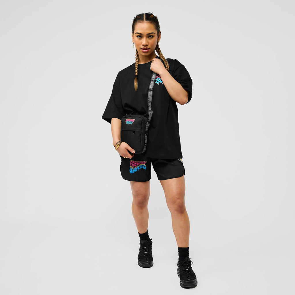 A model wearing a black oversized t-shirt, black shorts, and black boots, styled with a black crossbody bag featuring colorful graphics, set against a plain light gray studio background.