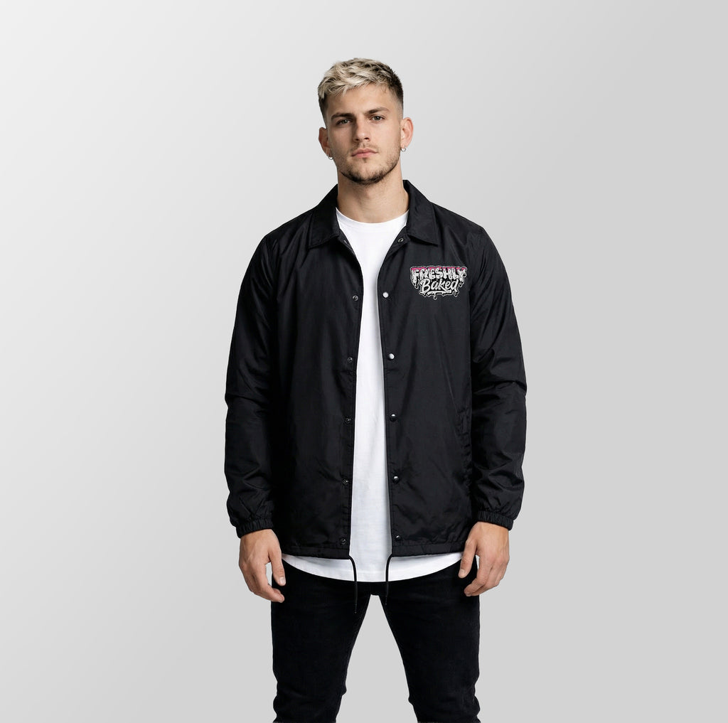 A man wearing a black coach jacket with a graphic print on the left chest over a white t-shirt and black pants, standing against a light gray studio background.