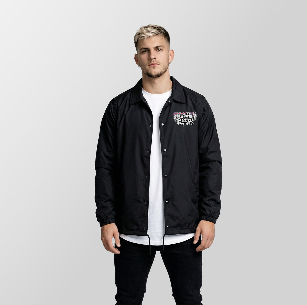 A man wearing a black coach jacket with a graphic print on the left chest over a white t-shirt and black pants, standing against a light gray studio background.