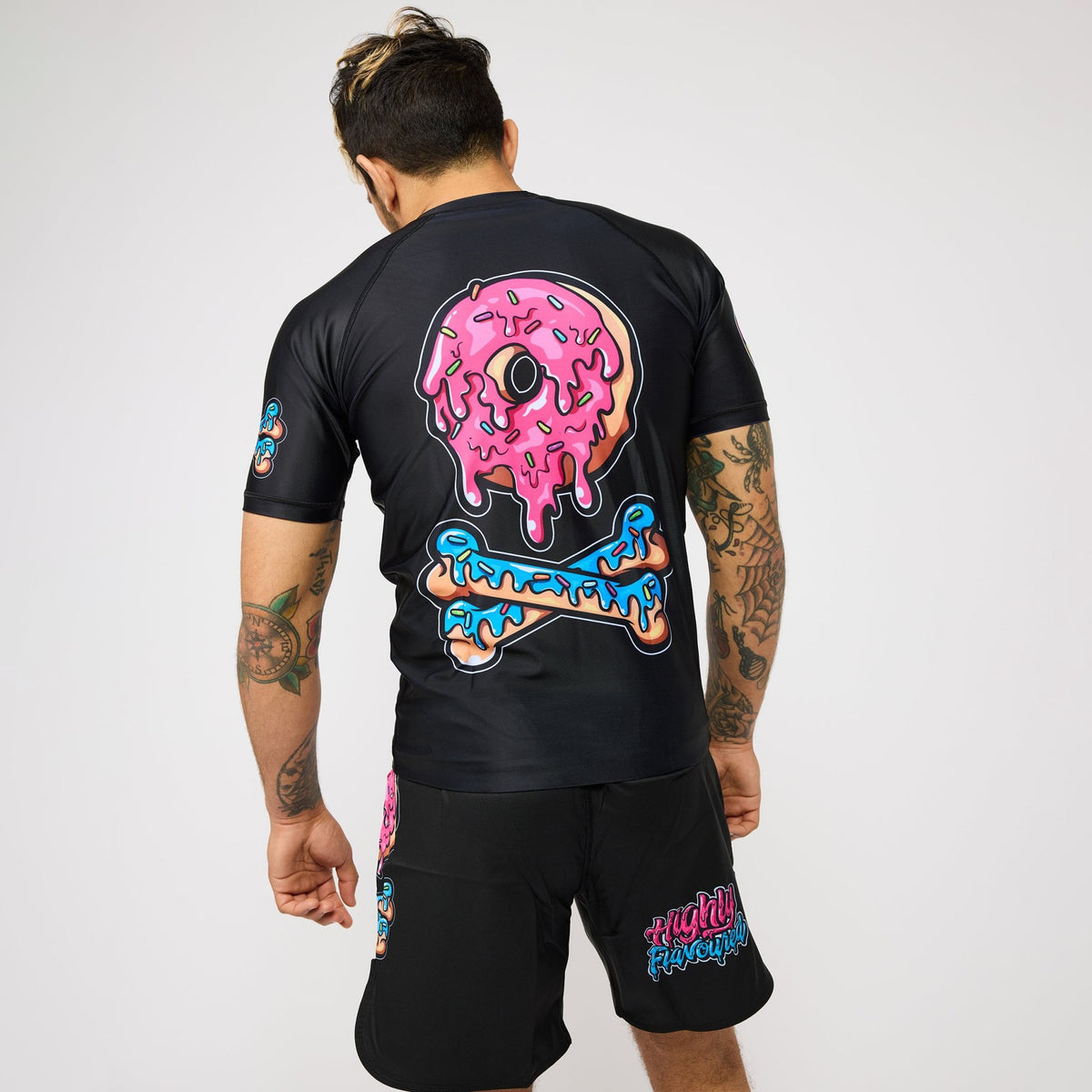 A man shown from the back wearing a black rash guard and matching black shorts with a large pink donut graphic and blue  text on the back, against a light gray studio background.