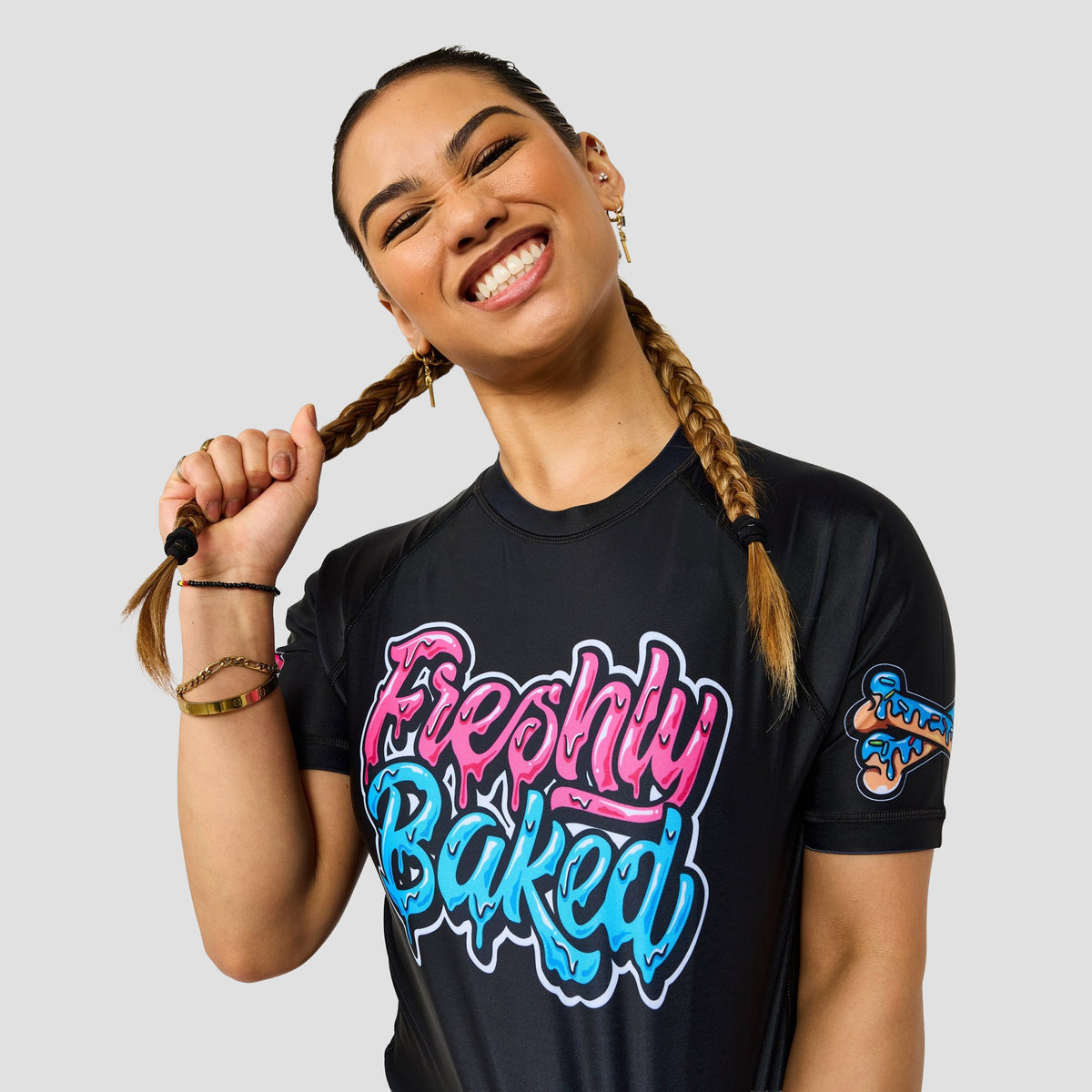 A smiling woman with braided hair wears a black graphic T-shirt with 'Freshly Baked' in pink and blue bubble letters on the front, plus a small 'Freshly Baked' logo on the sleeve, against a light gray background.