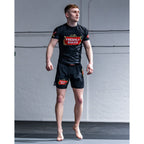 A man wearing a black athletic rash guard and matching black shorts with red and gold 'Freshly Baked' logos, standing barefoot on a mat in front of a white wall.