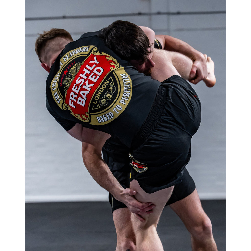 Two people grappling in a gym, with the person on top wearing black shorts and a black top featuring a large red, gold, and white circular graphic on the back that reads 'Freshly Baked London'.