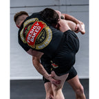 Two people grappling in a gym, with the person on top wearing black shorts and a black top featuring a large red, gold, and white circular graphic on the back that reads 'Freshly Baked London'.
