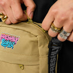 Close-up of a person's hands with rings opening the zipper of a tan canvas crossbody bag. The bag features a colorful 'Freshly Baked' graffiti patch on the front pocket and a black webbed strap with the brand name printed vertically.