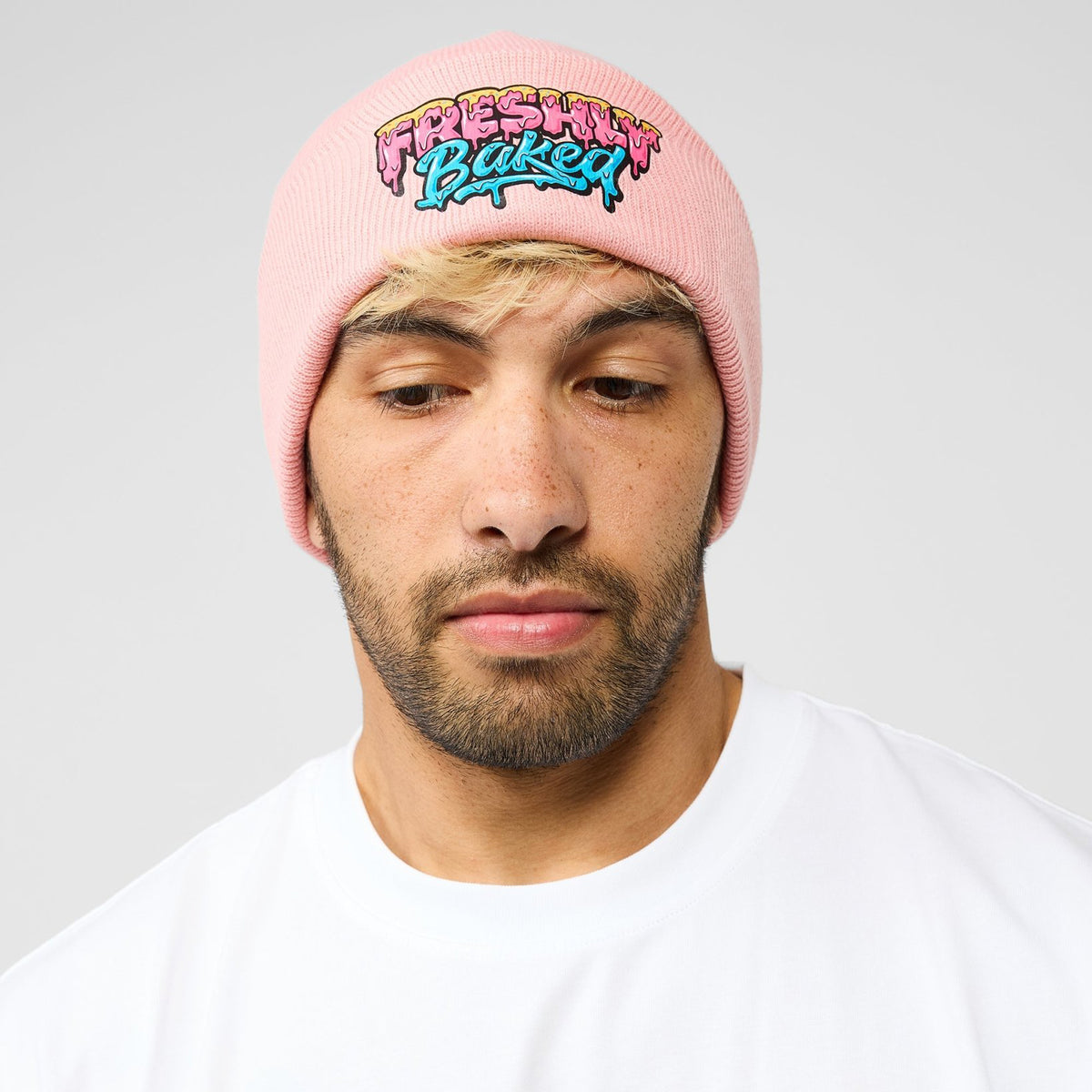 A man wearing a light pink knit beanie with a colorful embroidered graphic that reads 'FRESHLY BUCKED' in a graffiti-style font, paired with a white t-shirt against a light gray background.