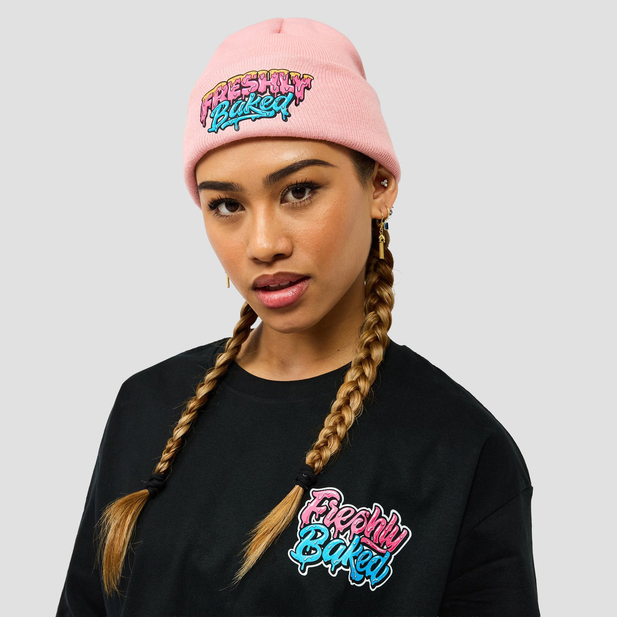 A model wearing a light pink beanie and a black sweatshirt, both featuring a colorful 'Freshly Baked' graphic logo on the front, set against a solid light gray background.