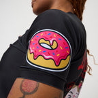 Close-up of a person wearing a black short-sleeve top with a large pink donut graphic on the upper arm, shown against a light neutral background.