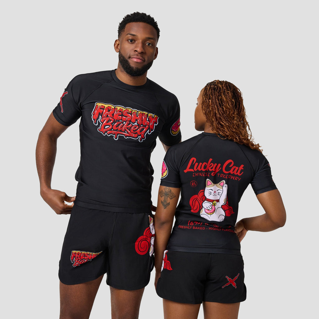 A man and a woman modeling matching black rash guard shirts and shorts with red and white graphics. The man shows the front of the shirt with 'Fresh Kid' text, and the woman shows the back of the shirt with a 'Lucky Cat' graphic.