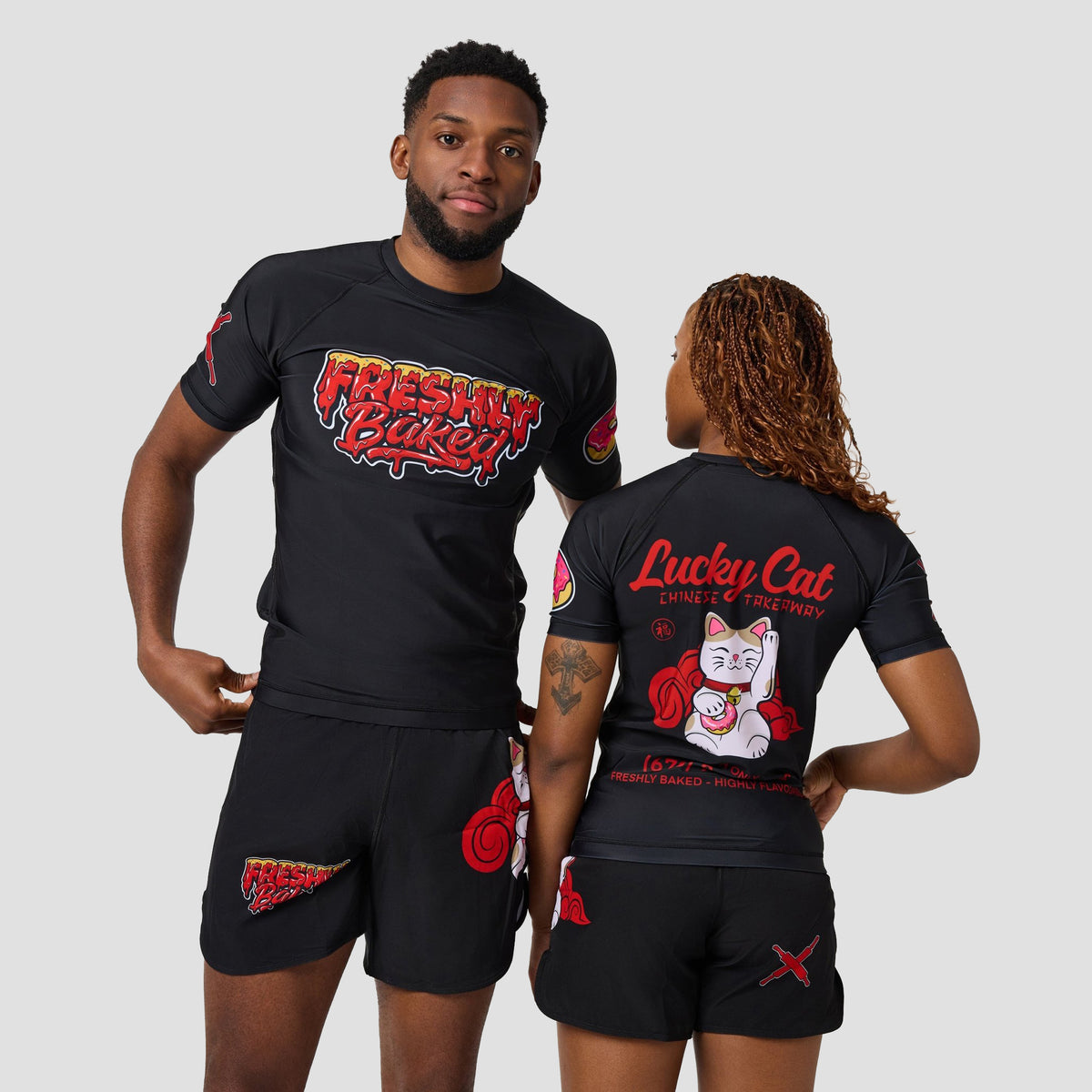 A man and a woman modeling matching black rash guard shirts and shorts with red and white graphics. The man shows the front of the shirt with 'Fresh Kid' text, and the woman shows the back of the shirt with a 'Lucky Cat' graphic.
