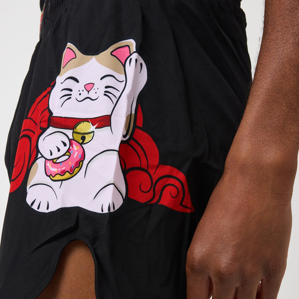 Close-up of a person wearing black shorts with a large printed cartoon cat graphic on the left leg. The cat is white with tan ears and paws, wearing a red collar with a gold bell and holding a pink donut, with red cloud-like accents.