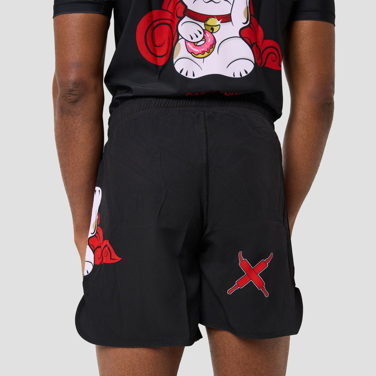 Black athletic shorts with red and white graphic prints, shown on a model from the back. The shorts feature a red 'X' logo on the left leg and a white cartoon character with red swirls on the right leg.