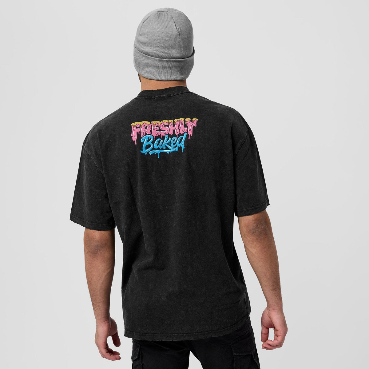 A man wearing a black washed t-shirt from the back, featuring a colorful 'Freshly Baked' graphic in pink, yellow, and blue on the upper back. He is also wearing a gray beanie, photographed against a solid light gray background.
