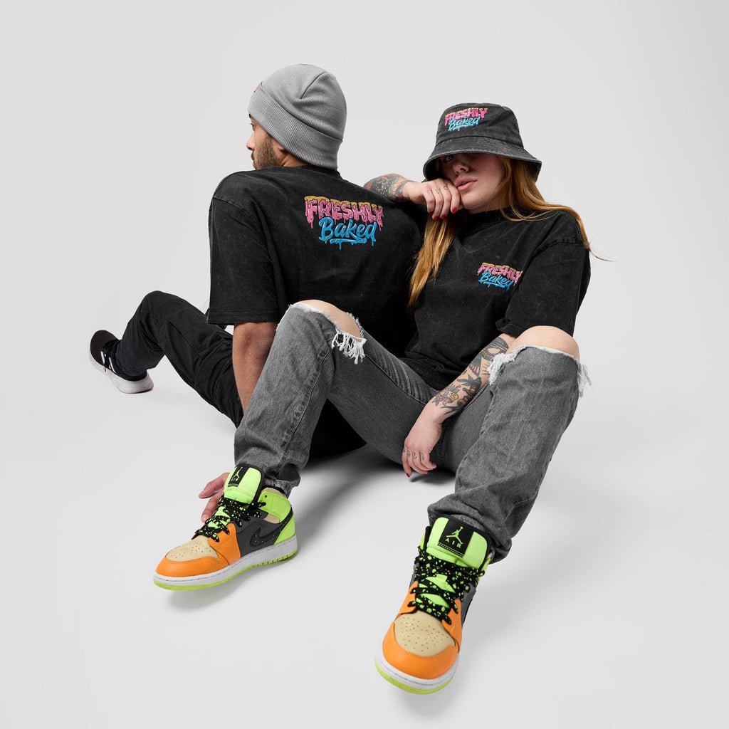 Two models sitting together wearing matching black graphic t-shirts with 'Freshly Baked' printed on the back and chest, paired with ripped gray jeans and bright orange, green, and black sneakers, against a light gray background.