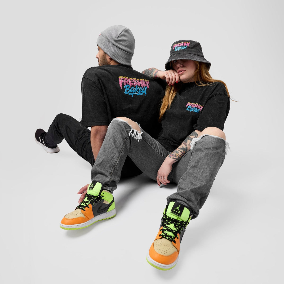 Two models sitting together wearing matching black graphic t-shirts with 'Freshly Baked' printed on the back and chest, paired with ripped gray jeans and bright orange, green, and black sneakers, against a light gray background.