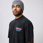 A man wearing a gray beanie and a faded black t-shirt, both featuring the 'Freshly Baked' logo in pink, yellow, and blue graffiti-style lettering, photographed against a light gray studio background.