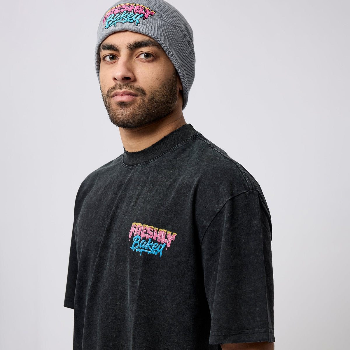 A man wearing a gray beanie and a faded black t-shirt, both featuring the 'Freshly Baked' logo in pink, yellow, and blue graffiti-style lettering, photographed against a light gray studio background.